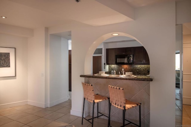 bar featuring light tile patterned flooring, decorative backsplash, dark brown cabinetry, and dark stone counters