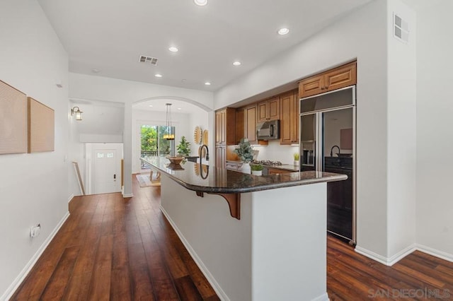 kitchen featuring built in refrigerator, dark hardwood / wood-style floors, a kitchen island with sink, a kitchen breakfast bar, and pendant lighting