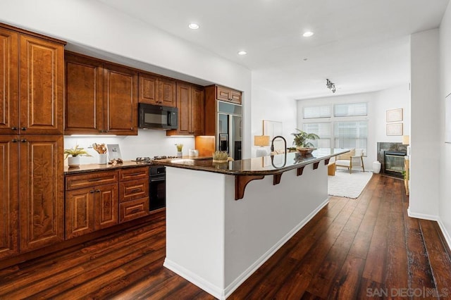 kitchen featuring a center island with sink, dark hardwood / wood-style floors, dark stone counters, a breakfast bar, and black appliances