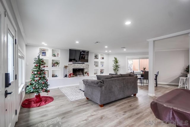 living room with built in features, a stone fireplace, crown molding, and light hardwood / wood-style flooring