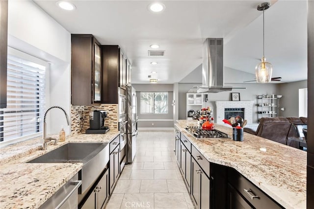 kitchen with light stone countertops, pendant lighting, island range hood, stainless steel gas stovetop, and dark brown cabinets