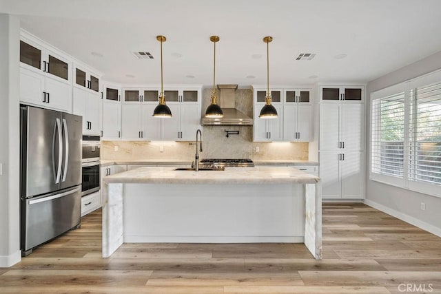 kitchen with decorative light fixtures, wall chimney range hood, a center island with sink, sink, and stainless steel appliances