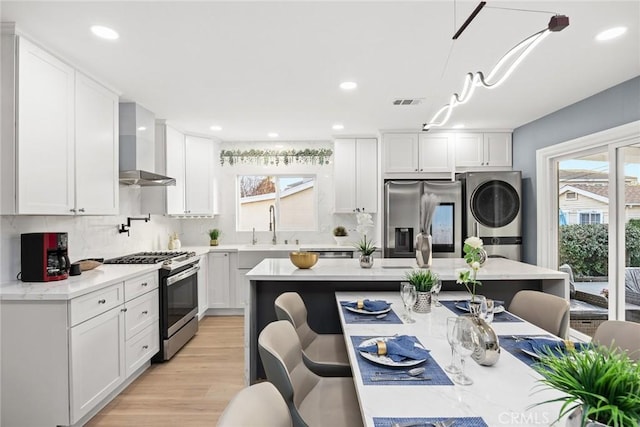 kitchen with stacked washer / drying machine, light hardwood / wood-style floors, stainless steel appliances, wall chimney range hood, and white cabinets