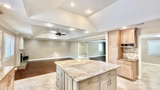 kitchen featuring light stone countertops, a kitchen island, light brown cabinets, a raised ceiling, and crown molding