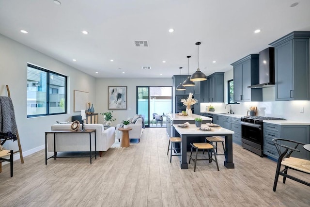 kitchen featuring wall chimney range hood, stainless steel gas range oven, light hardwood / wood-style flooring, hanging light fixtures, and a kitchen breakfast bar