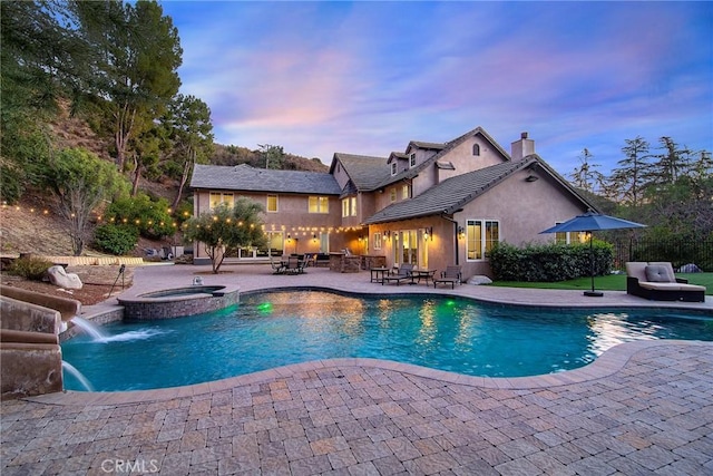 pool at dusk with pool water feature, an in ground hot tub, and a patio