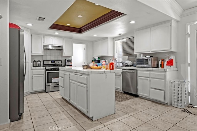 kitchen featuring appliances with stainless steel finishes, tile counters, a kitchen island, white cabinetry, and a tray ceiling