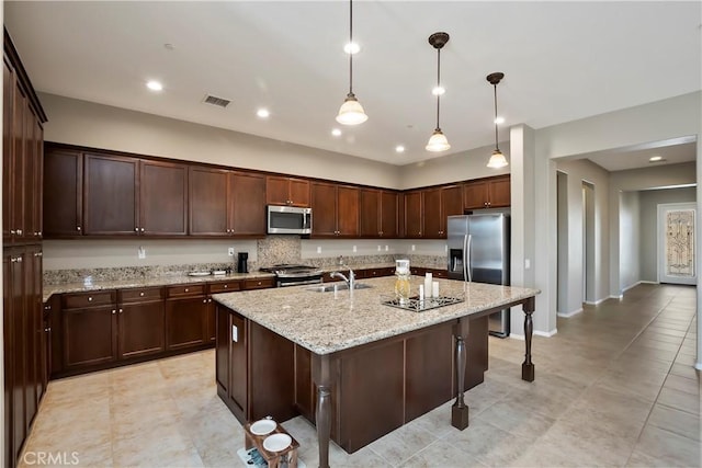 kitchen featuring a center island with sink, sink, hanging light fixtures, light stone countertops, and appliances with stainless steel finishes