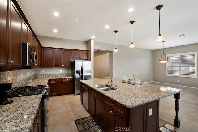 kitchen featuring an island with sink, appliances with stainless steel finishes, decorative light fixtures, light stone counters, and sink