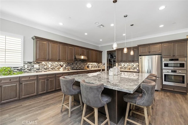 kitchen featuring pendant lighting, appliances with stainless steel finishes, dark brown cabinets, and an island with sink