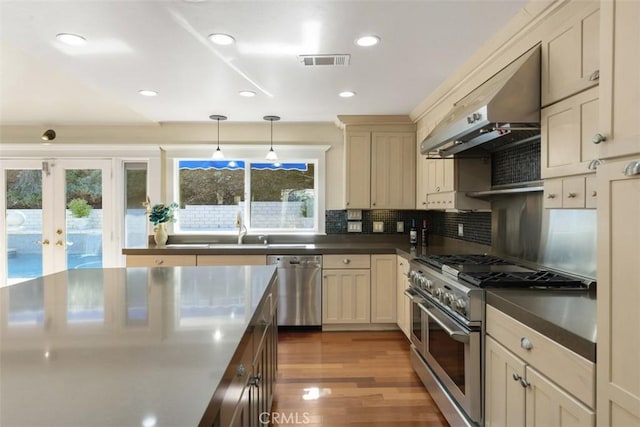 kitchen featuring sink, appliances with stainless steel finishes, pendant lighting, cream cabinets, and wall chimney range hood