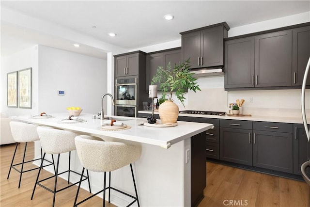 kitchen with stainless steel double oven, dark hardwood / wood-style flooring, white gas stovetop, and an island with sink