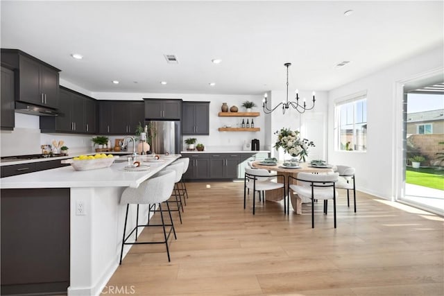 kitchen featuring an inviting chandelier, a center island with sink, stainless steel fridge, decorative light fixtures, and light hardwood / wood-style flooring