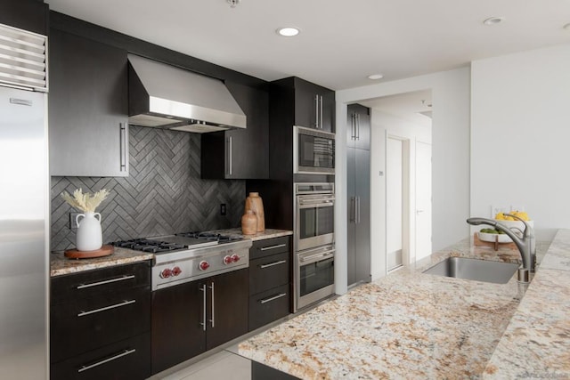 kitchen featuring wall chimney range hood, backsplash, built in appliances, light stone counters, and sink