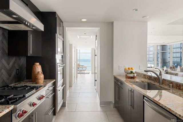 kitchen with light stone countertops, appliances with stainless steel finishes, wall chimney range hood, sink, and light tile patterned flooring