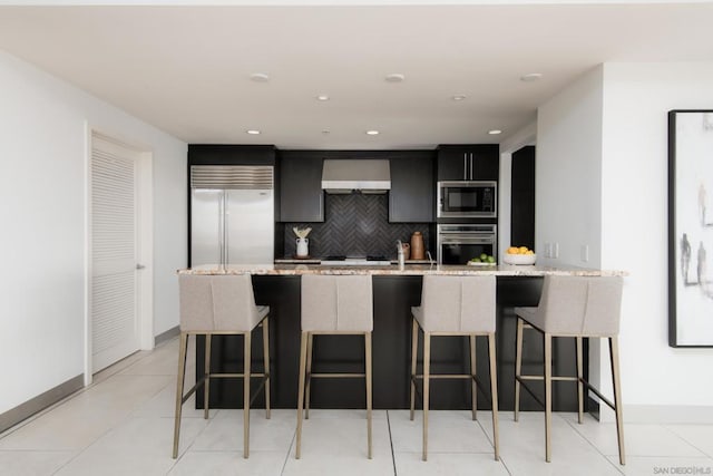 kitchen featuring light stone counters, backsplash, a center island with sink, and built in appliances