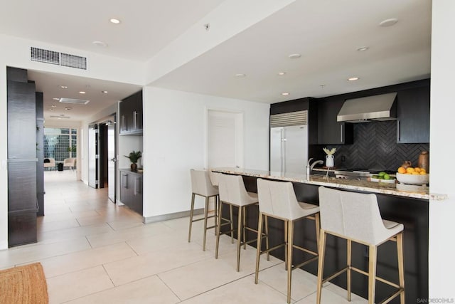 kitchen featuring light stone counters, wall chimney exhaust hood, stainless steel built in fridge, and tasteful backsplash