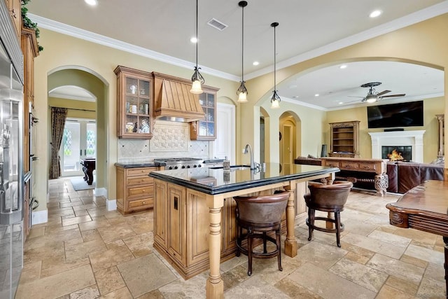 kitchen with ceiling fan, an island with sink, decorative backsplash, premium range hood, and pendant lighting