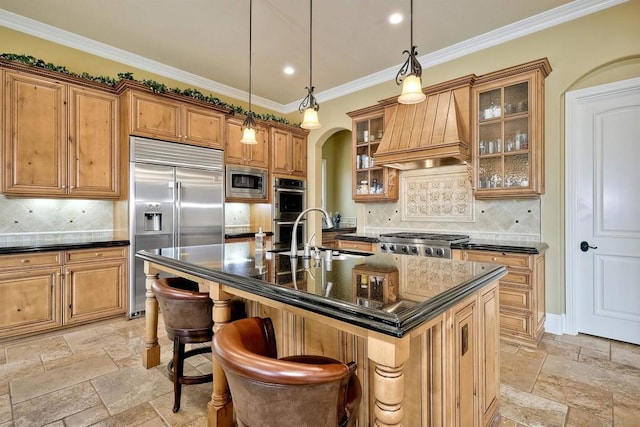 kitchen featuring a center island with sink, built in appliances, premium range hood, sink, and tasteful backsplash