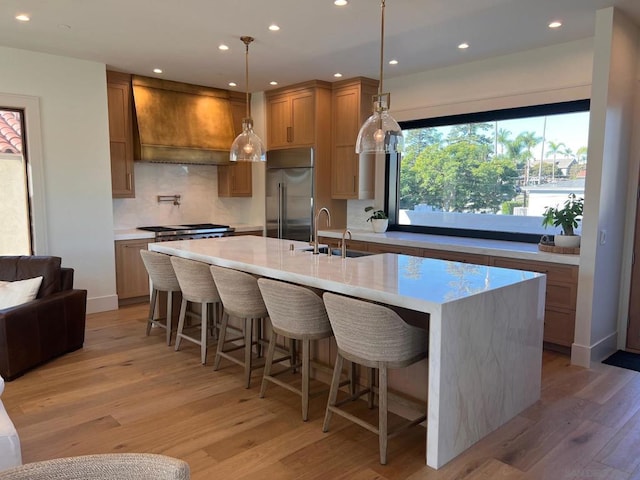 kitchen with sink, custom range hood, an island with sink, a kitchen bar, and built in fridge