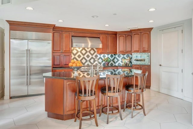 kitchen with backsplash, extractor fan, built in appliances, and a center island with sink