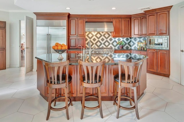 kitchen featuring ventilation hood, a kitchen island with sink, a kitchen breakfast bar, and built in appliances