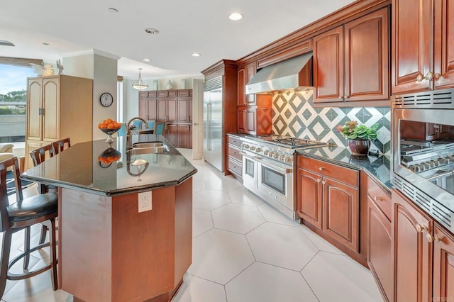 kitchen with wall chimney range hood, high end appliances, sink, backsplash, and a center island with sink