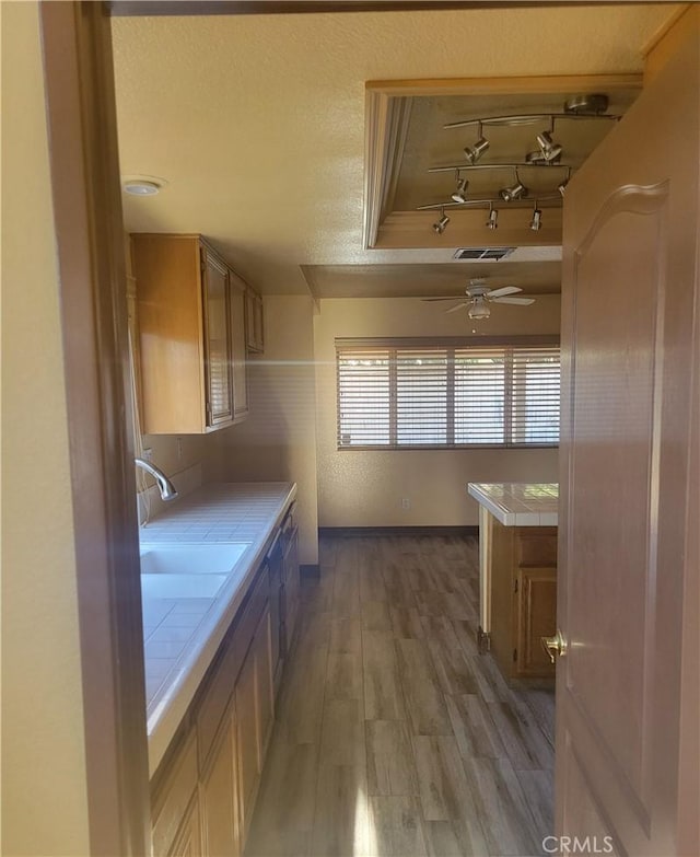 kitchen featuring ceiling fan, sink, a tray ceiling, light wood-type flooring, and tile counters