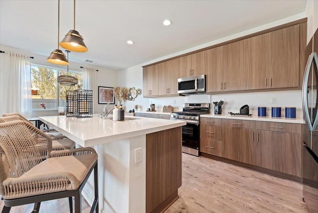 kitchen featuring appliances with stainless steel finishes, an island with sink, hanging light fixtures, light wood-type flooring, and a breakfast bar area