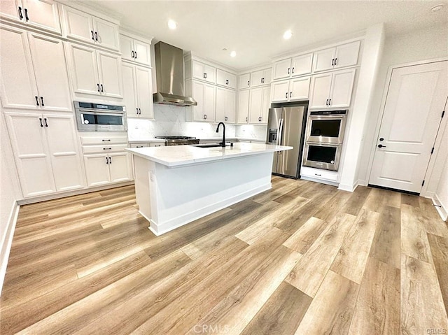 kitchen featuring a center island with sink, appliances with stainless steel finishes, wall chimney range hood, white cabinets, and sink