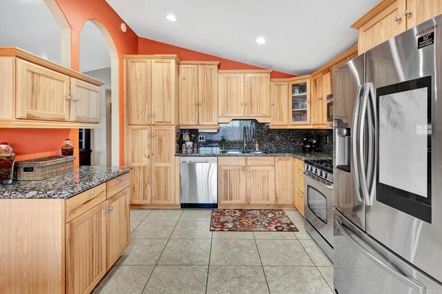 kitchen featuring light brown cabinetry, stainless steel appliances, dark stone counters, and vaulted ceiling