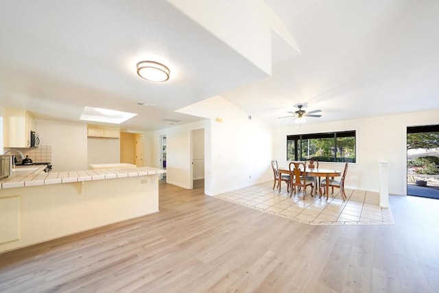 kitchen with a breakfast bar area, backsplash, tile counters, ceiling fan, and light hardwood / wood-style flooring