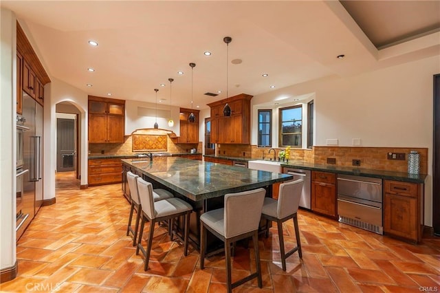 kitchen with pendant lighting, stainless steel appliances, dark stone countertops, an island with sink, and premium range hood