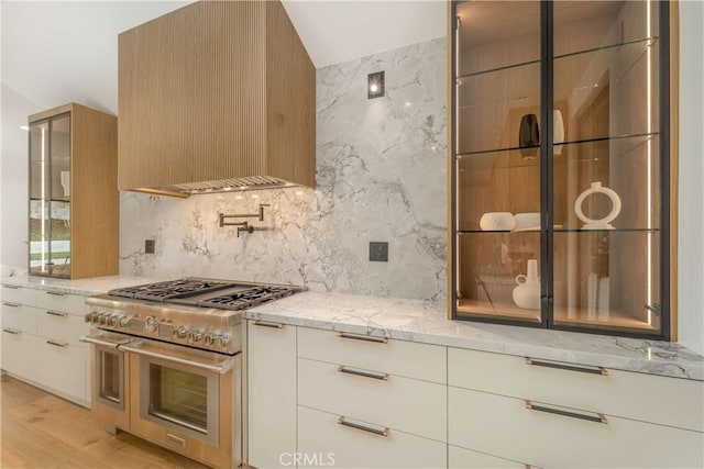 kitchen with tasteful backsplash, white cabinets, light stone counters, and high end range