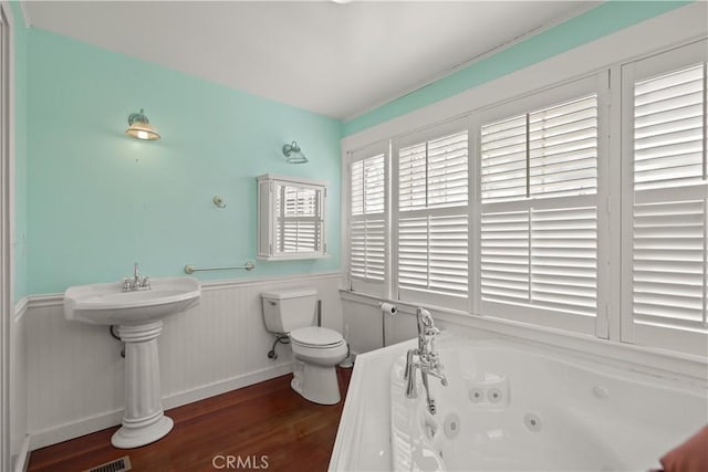 full bath featuring a jetted tub, toilet, wood finished floors, and wainscoting