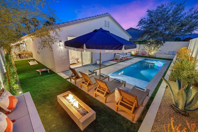 pool at dusk with a patio area, a fire pit, and a lawn