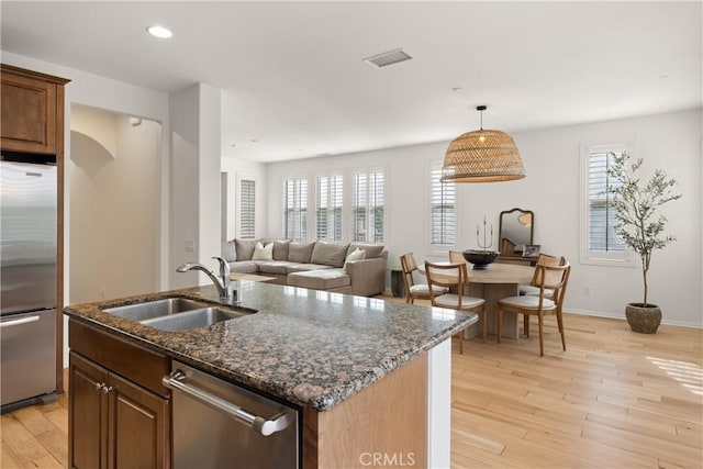 kitchen featuring appliances with stainless steel finishes, a kitchen island with sink, light wood-type flooring, dark stone counters, and sink