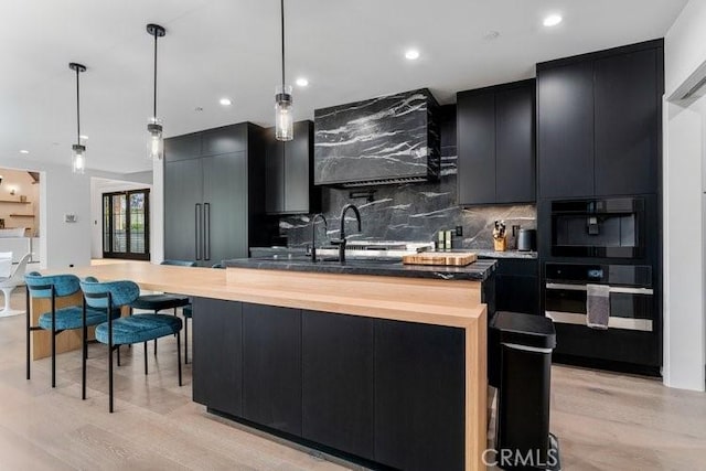 kitchen with backsplash, hanging light fixtures, a kitchen island with sink, oven, and light wood-type flooring