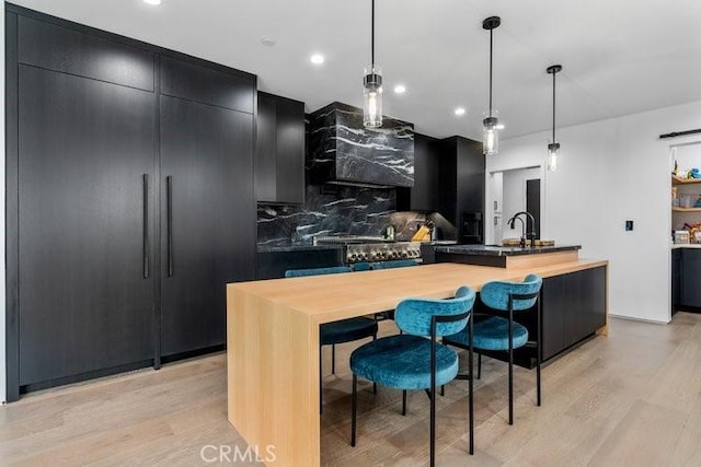 kitchen featuring decorative backsplash, decorative light fixtures, a kitchen island with sink, wall chimney exhaust hood, and light hardwood / wood-style flooring