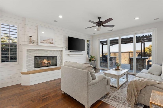 living room featuring ceiling fan, a tiled fireplace, dark wood-type flooring, and wood walls