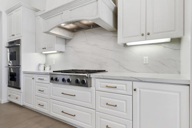 kitchen with custom exhaust hood, white cabinetry, light stone counters, appliances with stainless steel finishes, and backsplash