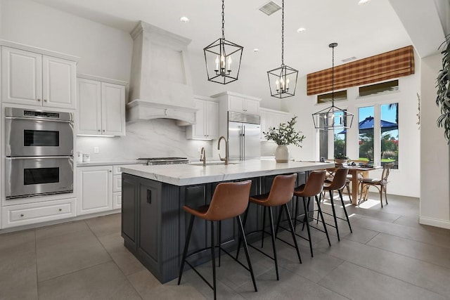 kitchen featuring a kitchen island with sink, white cabinetry, stainless steel appliances, decorative light fixtures, and custom exhaust hood