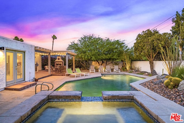 pool at dusk with an in ground hot tub, exterior fireplace, and a patio