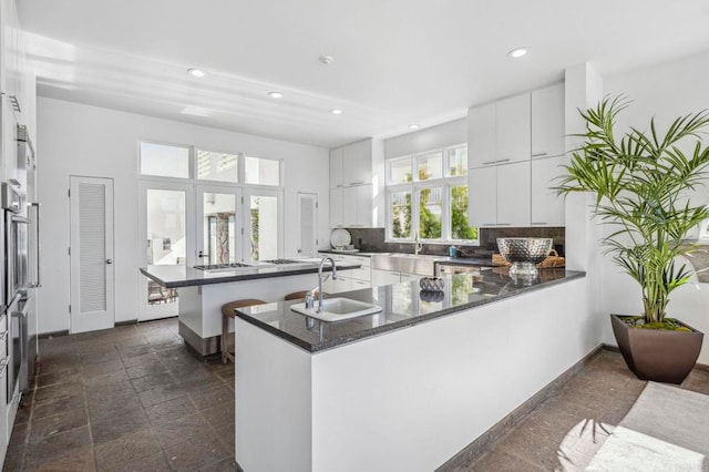 kitchen featuring kitchen peninsula, decorative backsplash, dark stone counters, white cabinets, and sink