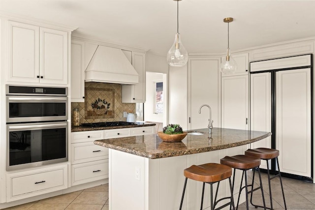 kitchen featuring appliances with stainless steel finishes, dark stone countertops, a kitchen island with sink, light tile patterned floors, and custom range hood