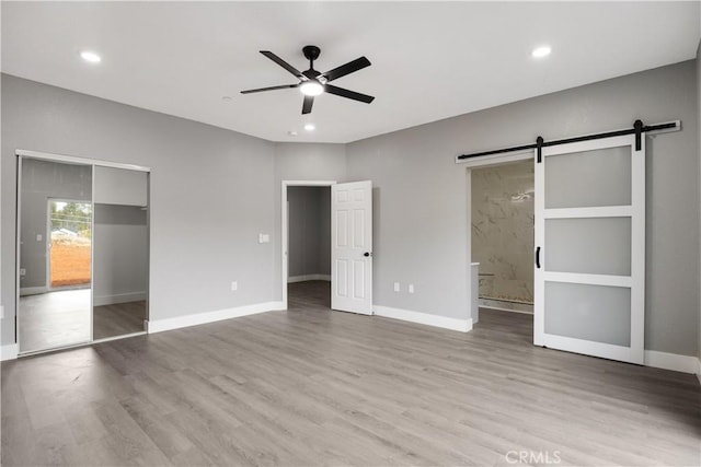 unfurnished bedroom with ensuite bathroom, a barn door, ceiling fan, and light wood-type flooring