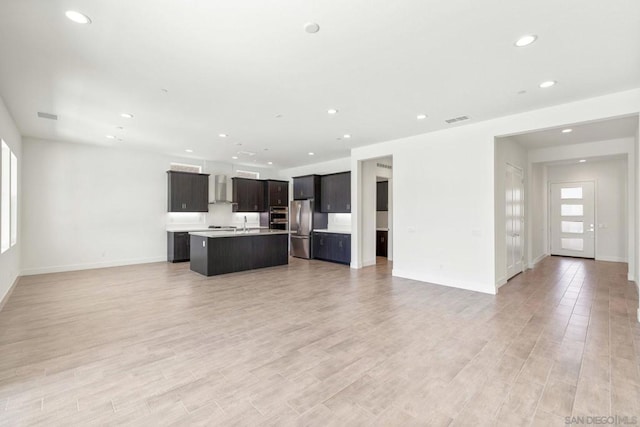 kitchen with wall chimney range hood, a kitchen island with sink, stainless steel appliances, and light wood-type flooring