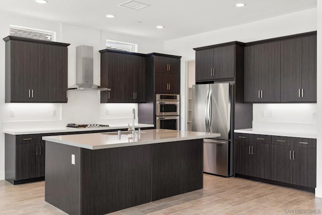 kitchen with stainless steel appliances, a kitchen island with sink, wall chimney exhaust hood, dark brown cabinetry, and sink