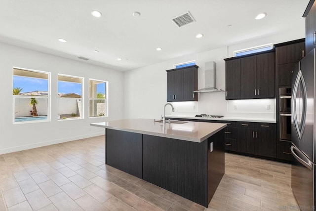 kitchen featuring sink, wall chimney range hood, appliances with stainless steel finishes, and a center island with sink