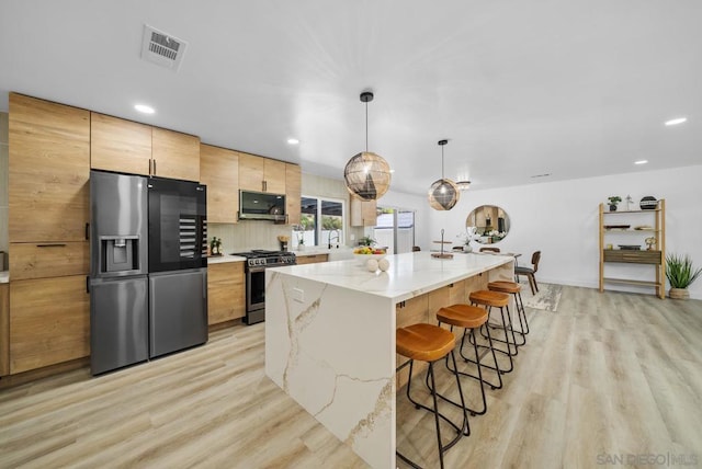 kitchen with stainless steel appliances, light stone countertops, a kitchen island, light brown cabinetry, and decorative light fixtures